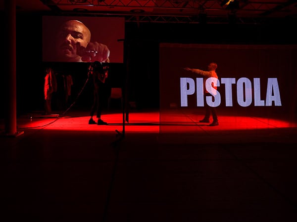 Ein Mann sitzt in der Mitte der Bühne und blickt in Richtung Publikum. Auf dieselbe Bühne wird das Gesicht eines anderen Mannes projiziert, so groß, dass es den ganzen Raum einnimmt.