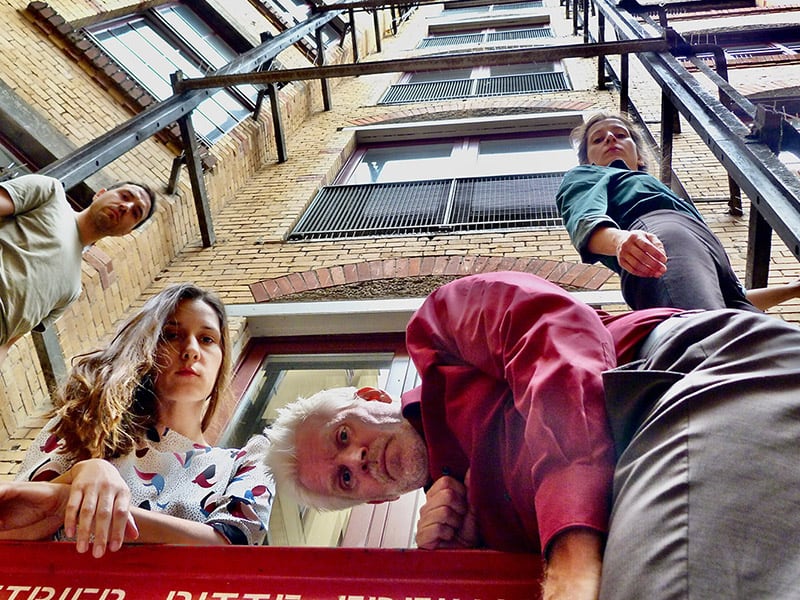 Photo in frog perspective of four people looking directly into the camera. They are in a backyard. In the background there are okka-colored bricks with a steel parapet. The two male and two female performers wear colorful everyday clothes.