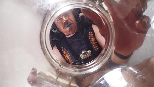 Close-up of a man with a red beard and blue lipstick in frog perspective, holding a transparent vessel into which he looks directly, at the same time looking into the camera.