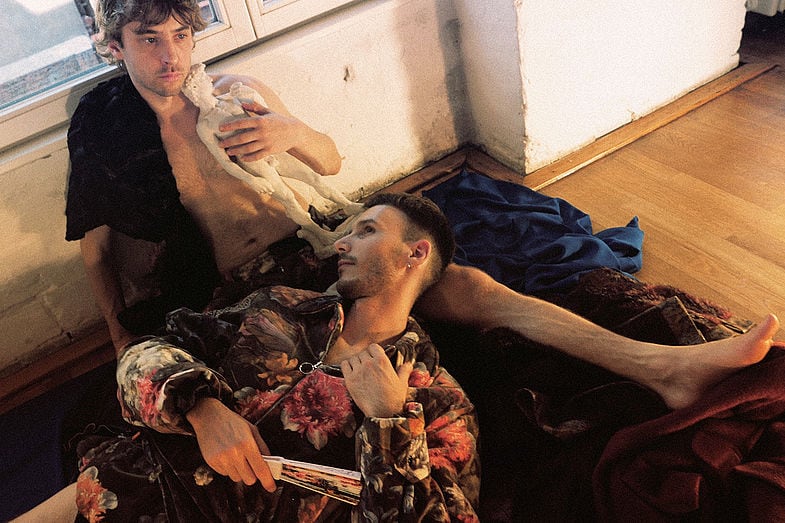 Photo of a man sitting on the wall, holding a small white sculpture as a doll, looking into the room, while another beds his head on his legs and holds a closed fan.