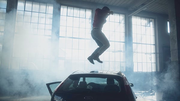 Ein dunkelblaues Auto in einer Industriehalle. Das Auto ist umgeben von weißem Rauch und steht im Gegenlicht einer Fensterfront. Auf dem Autodach springt eine Person in die Luft. Ihr Gesicht ist nicht zu erkennen. Sie trägt eine Jeans, eine rote Bomberjacke und Stiefel. Die Arme hat sie in die Luft erhoben.