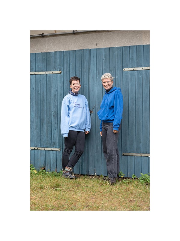 Two people are standing side by side in front of a blue wooden door on a lawn. Both are wearing blue sweaters, have short hair, and are white. They are smiling at the camera.