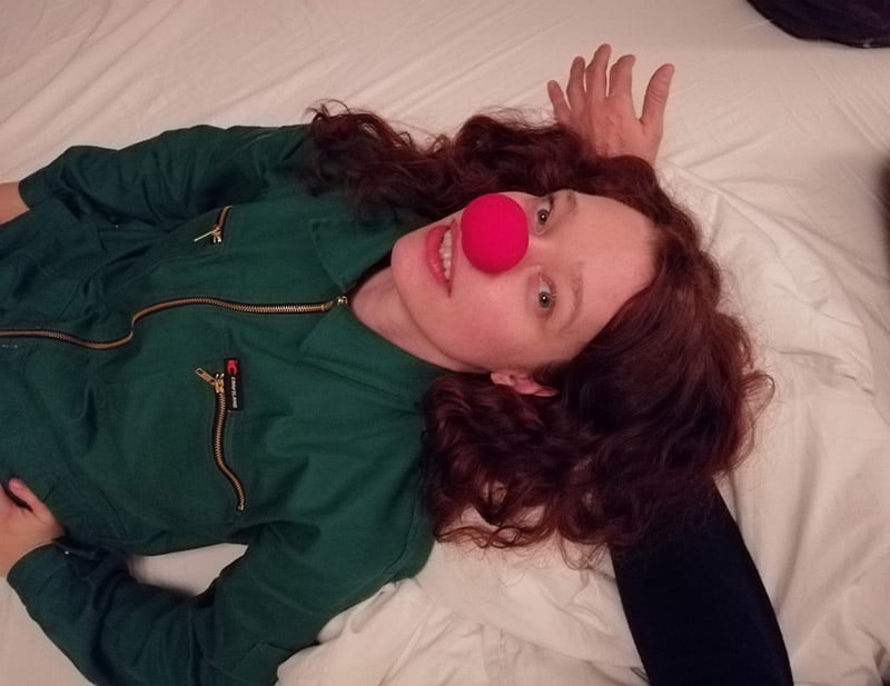Eine lächelnde, junge Frau in einem grünen Parka mit einer roten Clownsnase liegt auf einem weißen Tuch und schaut direkt in die Kamera, ihr Kopf ruht auf dem Arm einer weiteren Person, die nicht zu sehen ist.