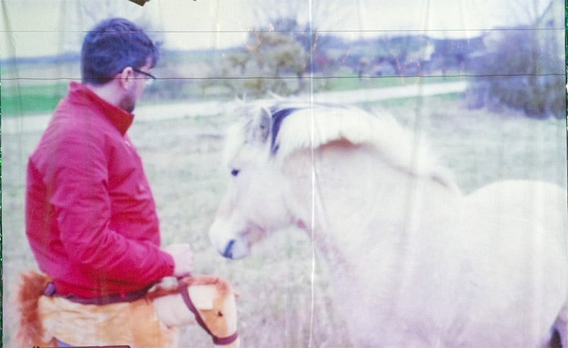 Image of a photo projected on an uneven canvas. Outside, a meadow and a path and trees in the background, in the foreground a pony and a middle-aged man with a pony costume around his hips, wearing a red jacket and black glasses.