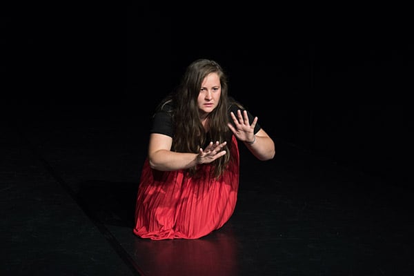 In the center of the photo is a young actress in a red shiny skirt kneeling in the black stage space, gesticulating, speaking towards the audience.