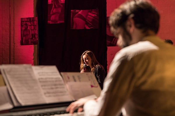 Photo of a man from behind at the piano out of focus in the foreground and a woman in the stage space sitting in the spotlight light speaking into a microphone, the wall is hung with nude photographs is bathed in a strong red light.