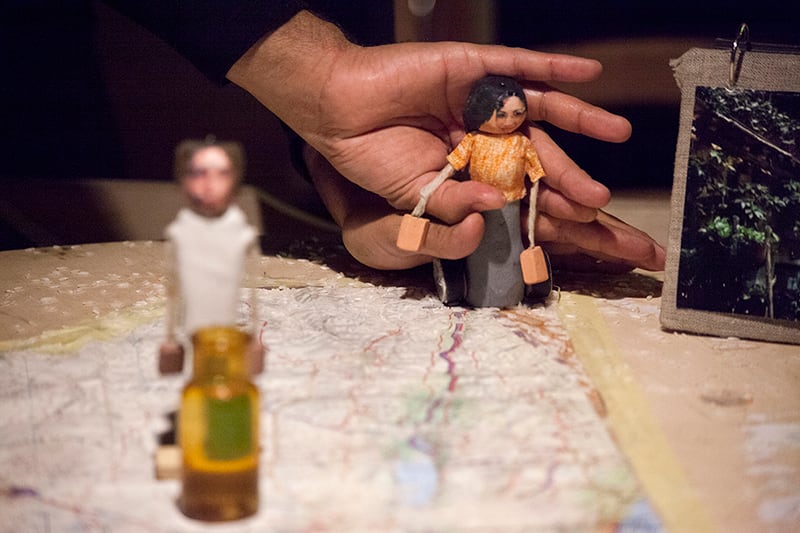 Closeup of three small puppets representing people, handled by two hands. The puppets are on top of a map on which rice grains have been scattered.