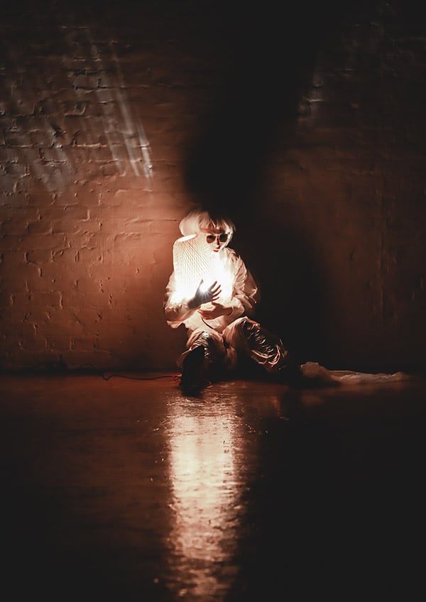 Photo of a white-clad performer wearing sunglasses in the black stage space illuminating herself with a flashlight, sitting against the back wall in the center of the image.