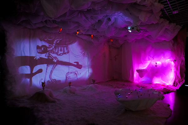 Eine immersive Kunstinstallation mit weißen Stoffen, die Wände, Decke und Boden bedecken, beleuchtet von violetten und pinken Lichtern. Ein Cartoonbild wird an eine Wand projiziert, während kleine bunte Lichter auf dem Boden leuchten.