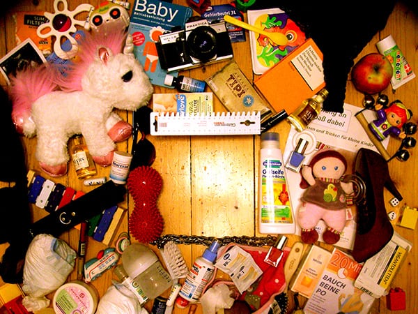 Photo of many scattered objects on a wooden floorboard: unicorn cuddly toy, camera, orange Reclam booklet, apple, tobacco and filter, children's toothpaste, colorful children's xylophone, dark red velvet lady's shoe with heel, tablet packaging, various guidebooks, two dice, Duplo bricks, small platic spoons, massage ball, glass and plastic bottles and flacons, other miscellaneous toys and hygiene articles.