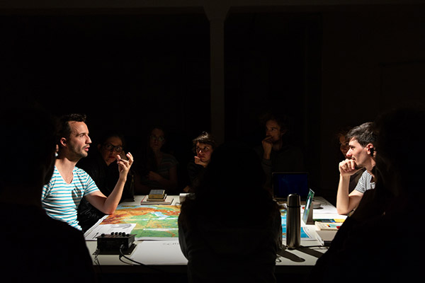 The performers in stage lighting are talking, sitting at a table, looking at a map. Books and booklets lie on the table. The audience is visible in the shadows.