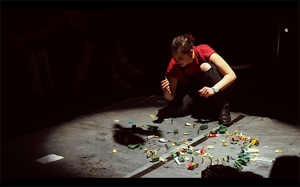 Female performer in black dungarees amongst plastic soldiers, empty food tins and garbage bags.